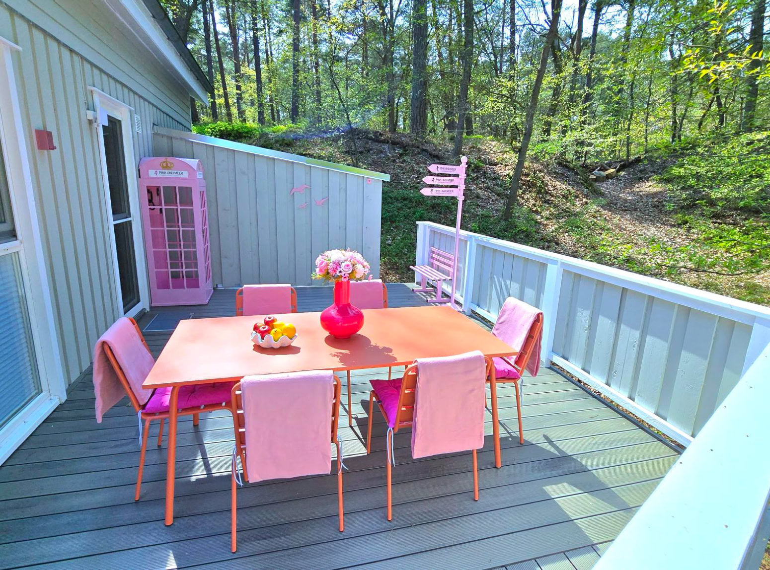 Terrasse mit einem Tisch und sechs rosa Stühlen, umgeben von einem weißen Geländer, neben einem hellblauen Gebäude. Im Hintergrund sind Bäume zu sehen.