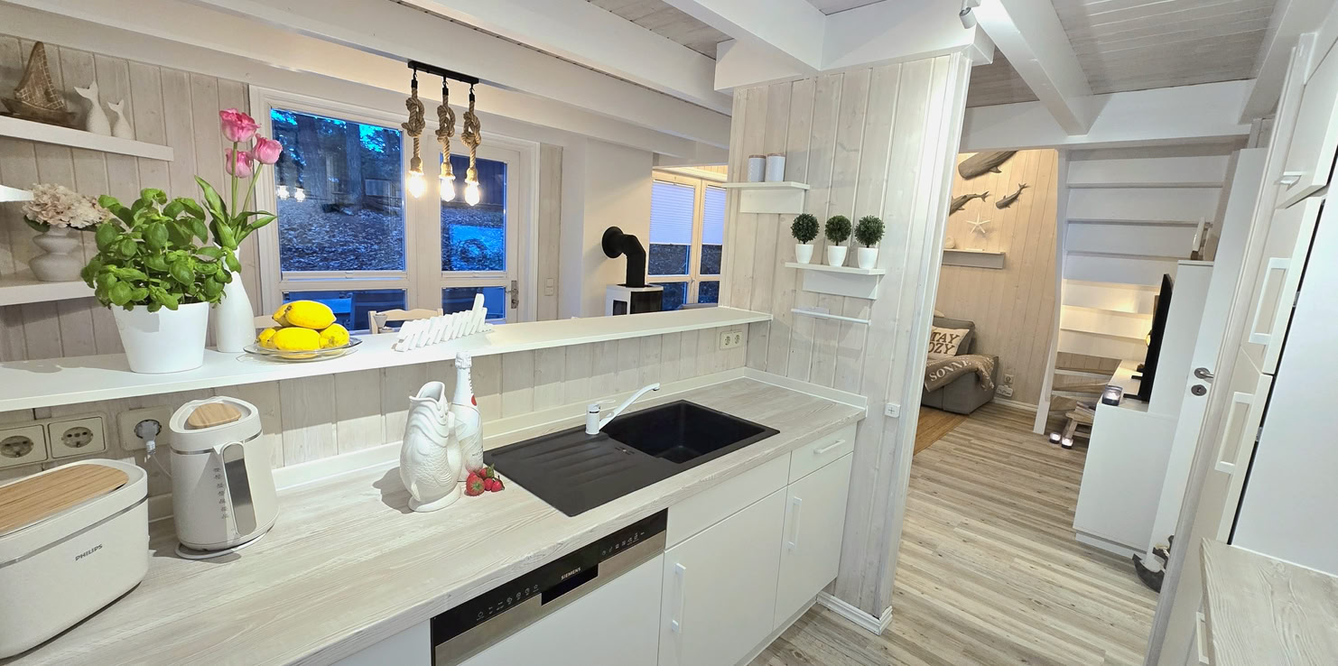 Kleiner Essbereich mit weißem Tisch und Stühlen, roten Sitzkissen, hölzernen Wänden und Decke, dekoriert mit Blumen und Geschirr, angrenzend an eine offene Küche.