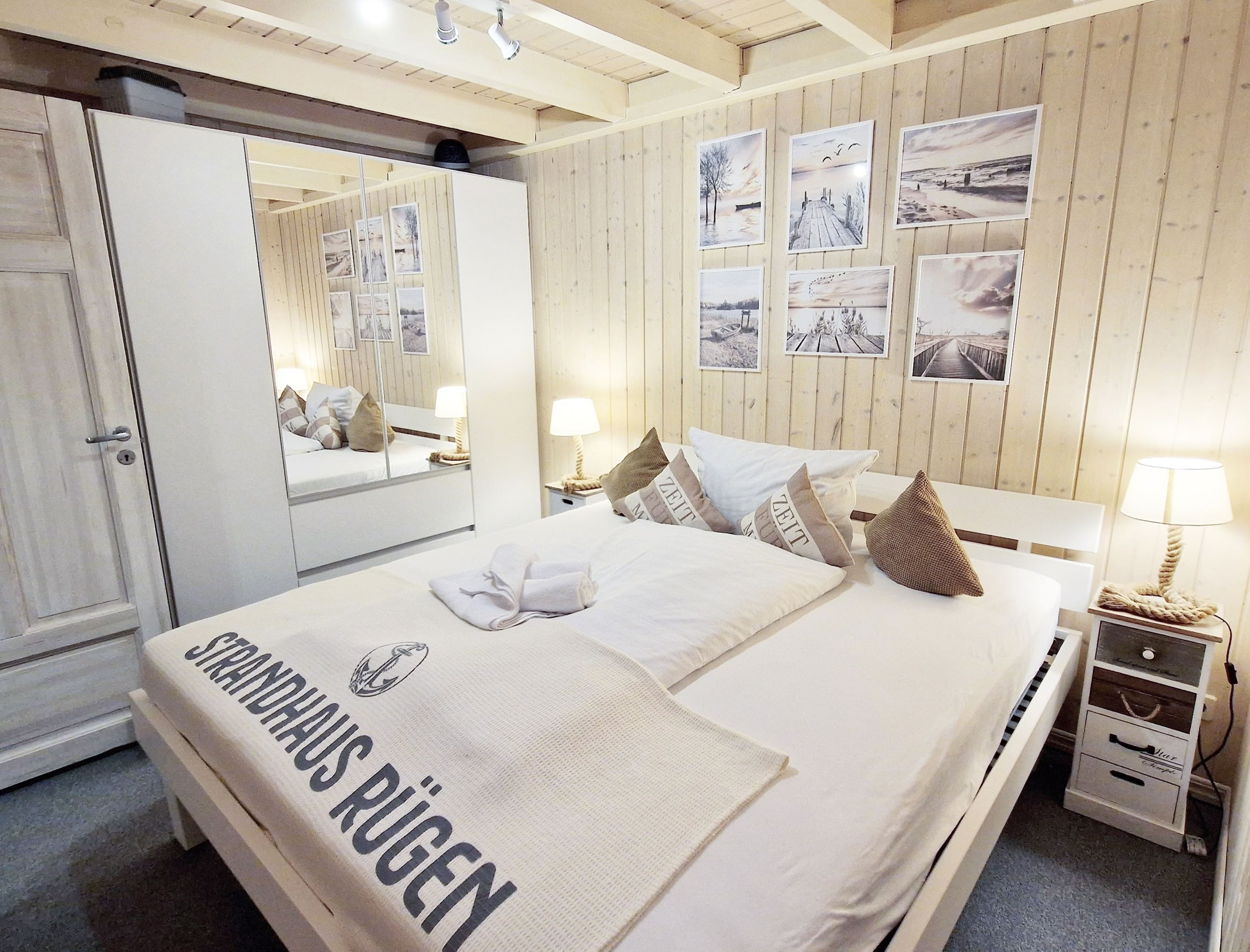 Ein Schlafzimmer mit einem Doppelbett, weißen Bettlaken und braunen Kissen, einem Kleiderschrank mit Spiegel, zwei Nachttischen mit Lampen und mehreren Bildern an der holzverkleideten Wand.