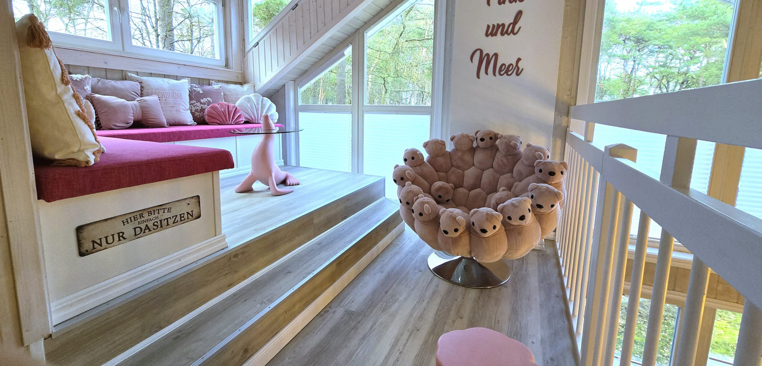 Gemütlicher Raum mit Dachschräge, rosa Sofa, rundem Sessel mit Plüschtieren, großen Fenstern und der Aufschrift "Pink and More" an der Wand.