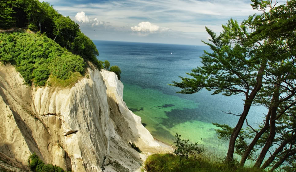 Kreidefelsen an der Küste mit grünem Wald oben, blauem Himmel und Meer im Hintergrund.
