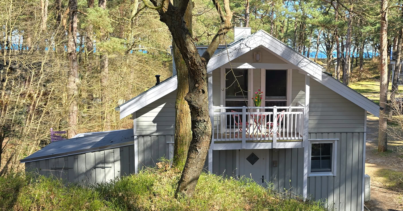 Graues Holzhaus mit Balkon, umgeben von Bäumen und Wald, bei sonnigem Wetter.
