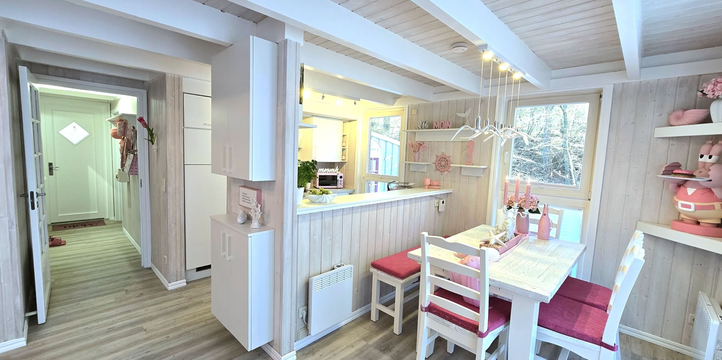 Kleiner Essbereich mit weißem Tisch und Stühlen, roten Sitzkissen, hölzernen Wänden und Decke, dekoriert mit Blumen und Geschirr, angrenzend an eine offene Küche.