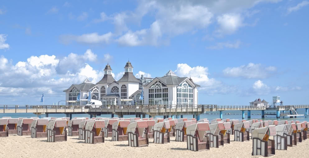 Strand mit vielen Strandkörben, im Hintergrund ein Gebäude auf einem Steg über dem Wasser, blauer Himmel mit einigen Wolken.