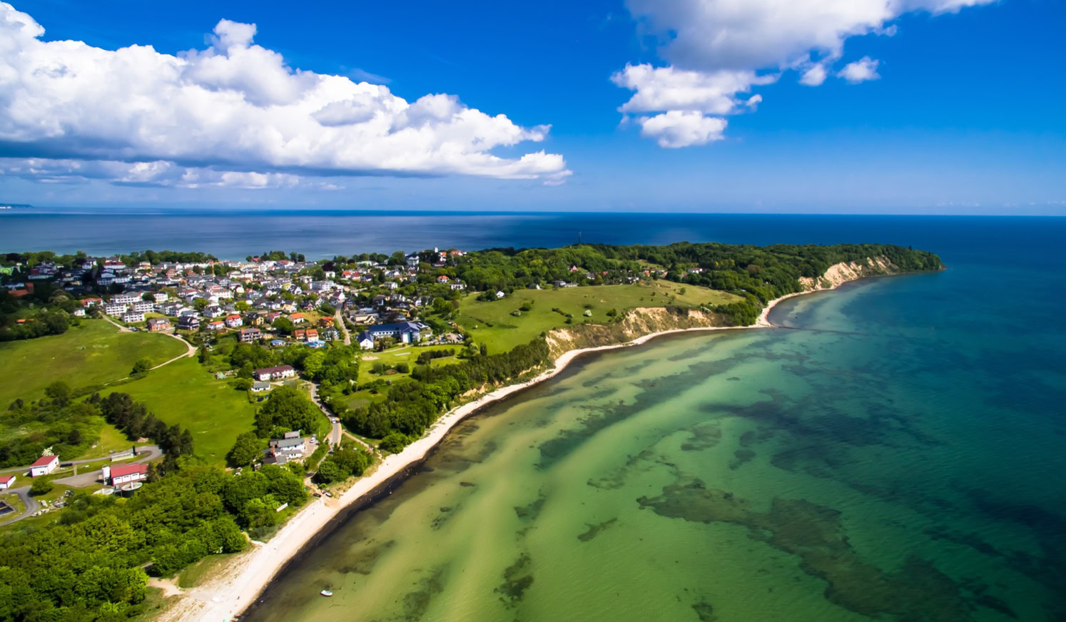 Luftaufnahme einer Küstenlandschaft mit klarem Wasser, Sandstrand, grünen Wiesen und einer Siedlung im Hintergrund unter einem blauen Himmel mit weißen Wolken.