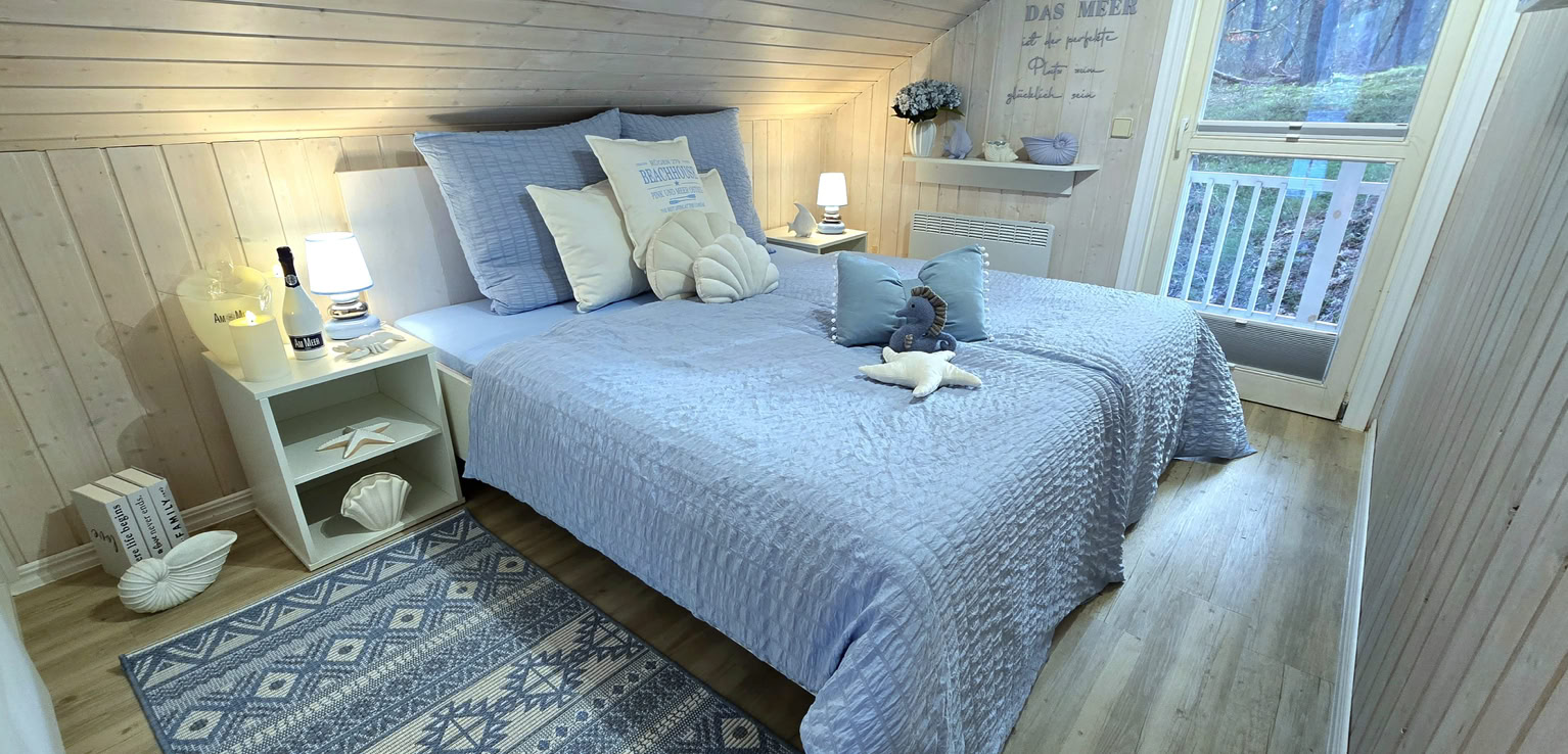 Gemütliches Schlafzimmer mit einem Doppelbett, blauen Bettdecken, mehreren Kissen, einem Nachttisch mit Lampe, einem Teppich und einem Fenster mit Blick nach draußen.