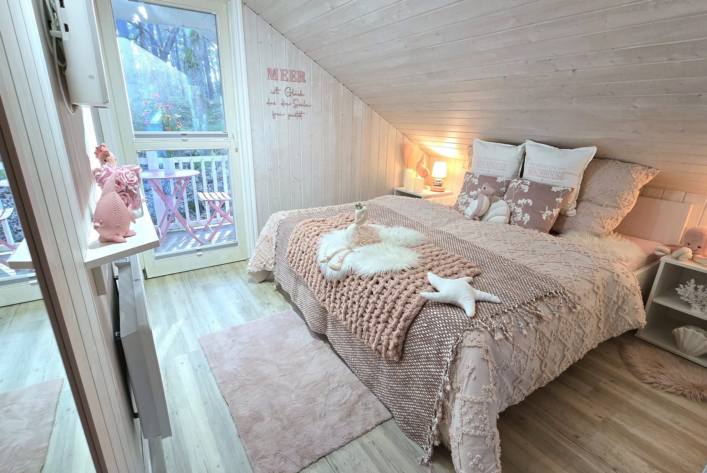 Gemütliches Schlafzimmer mit Dachschräge, Doppelbett mit mehreren Kissen, Nachttischlampen, Fenster mit Blick nach draußen und dekorativen Elementen.