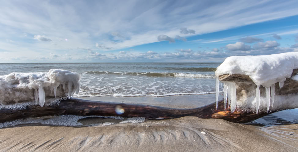 Verschneiter Strand mit gefrorenem Treibholz und Eiszapfen, im Hintergrund das Meer und ein bewölkter Himmel.