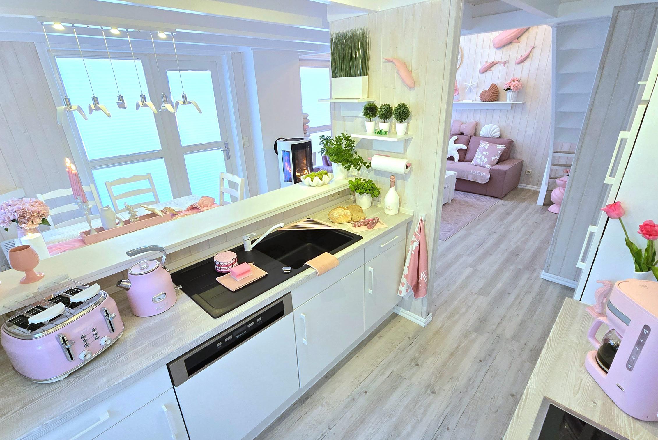 Helle Küche mit rosa Akzenten, einschließlich Toaster und Kaffeemaschine, weißen Schränken, einem Herd und einem Waschbecken. Im Hintergrund ist ein Wohnzimmer sichtbar.