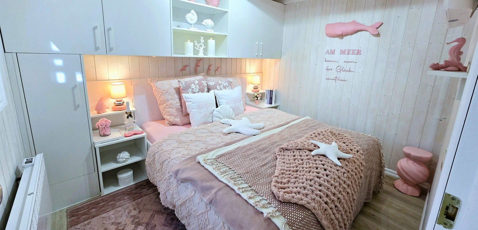 Gemütliches Schlafzimmer mit einem Doppelbett, dekoriert in Rosa- und Weißtönen, mit Kissen und Decken. An der Wand hängt ein Bild mit einem Wal. Regale mit Dekorationen befinden sich über dem Bett.