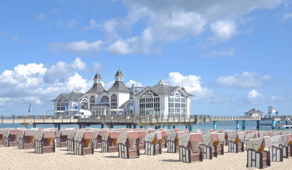 Strand mit Strandkörben im Vordergrund, dahinter ein Gebäude auf einer Seebrücke, blauer Himmel mit einigen Wolken.