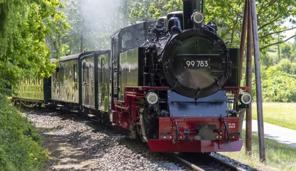 Dampflok mit Waggons fährt auf einem Gleis durch eine grüne Landschaft.