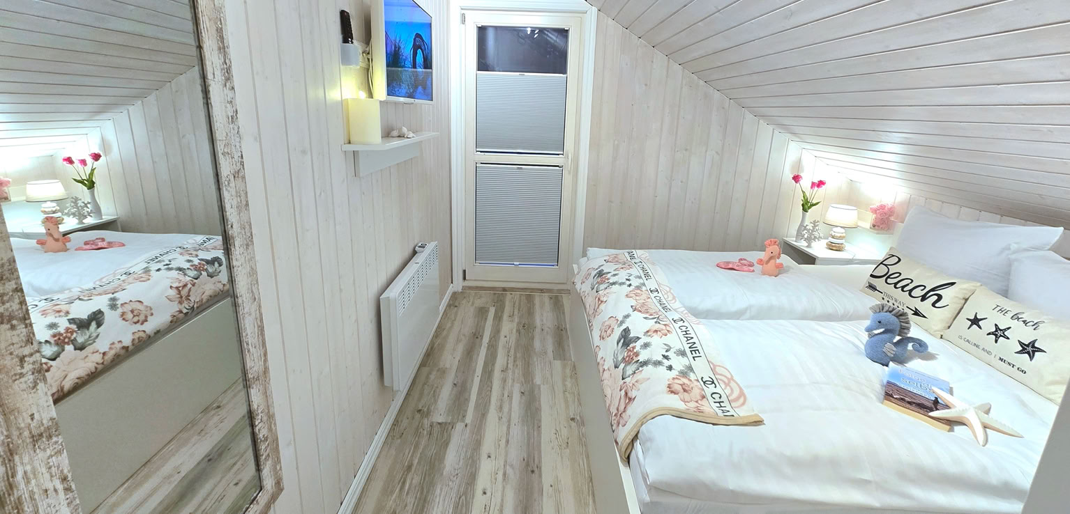 Gemütliches Schlafzimmer mit einem Doppelbett, dekoriert in Rosa- und Weißtönen, mit Kissen und Decken. An der Wand hängt ein Bild mit einem Wal. Regale mit Dekorationen befinden sich über dem Bett.