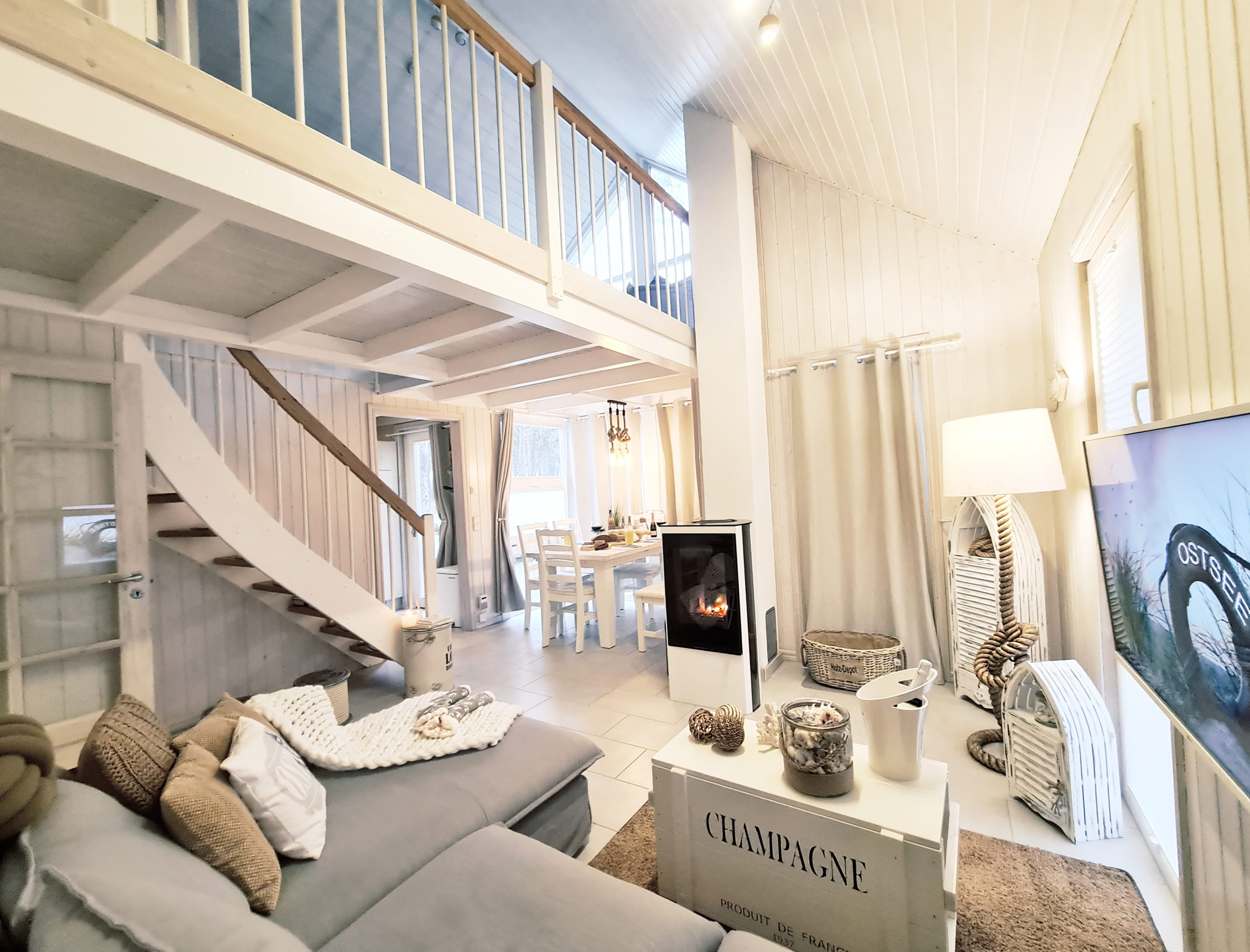 Gemütliches Wohnzimmer mit grauem Sofa, Kamin, großem Fernseher und offener Treppe zu einer Galerie. Helle, moderne Einrichtung mit Holzdetails und dekorativen Kissen.