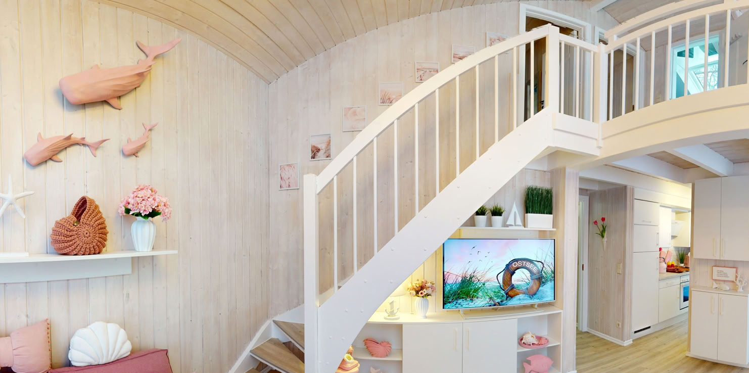 Ein gemütliches Wohnzimmer mit rosa Sofa, dekorativen Kissen, weißen Regalen mit rosa Dekorationen, einem Fernseher unter einer Treppe und Holzboden.