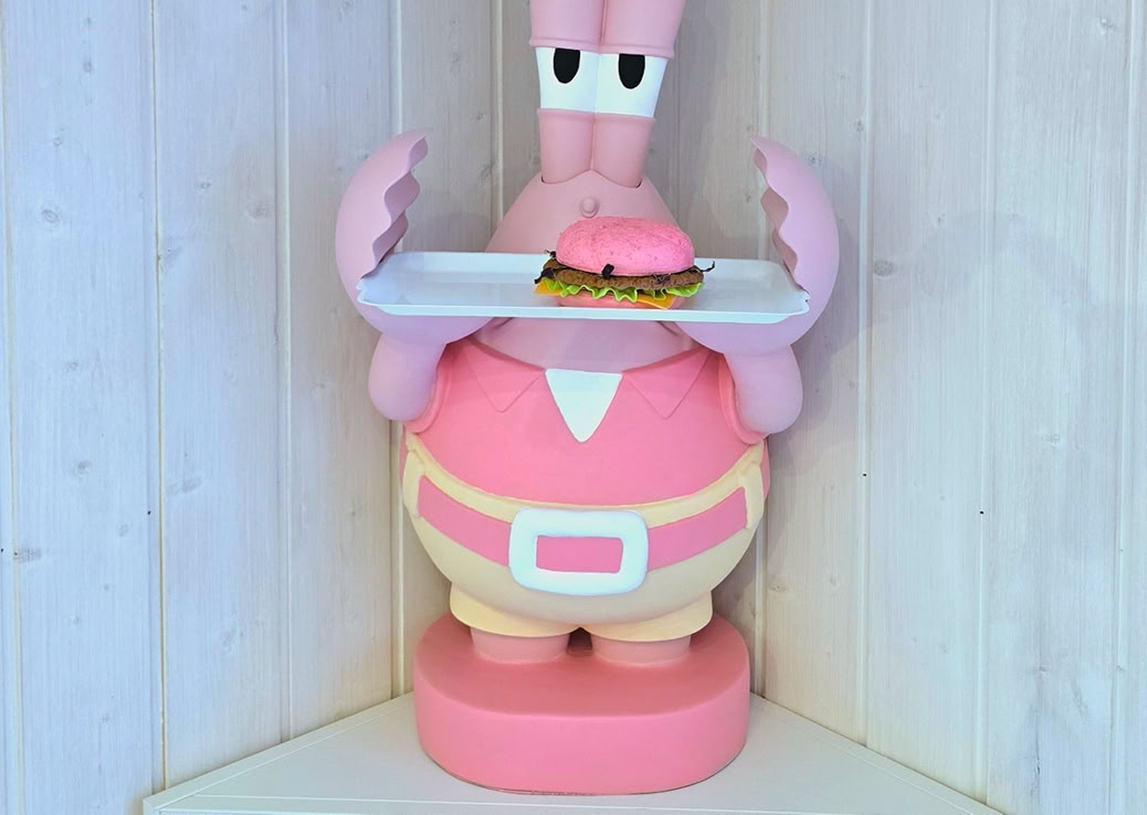 Eine pinke Cartoon-Figur mit einer Schürze hält ein Tablett mit einem Donut. Darüber befindet sich ein weißes Regal mit einer Vase voller rosa Blumen und einer Muschel.