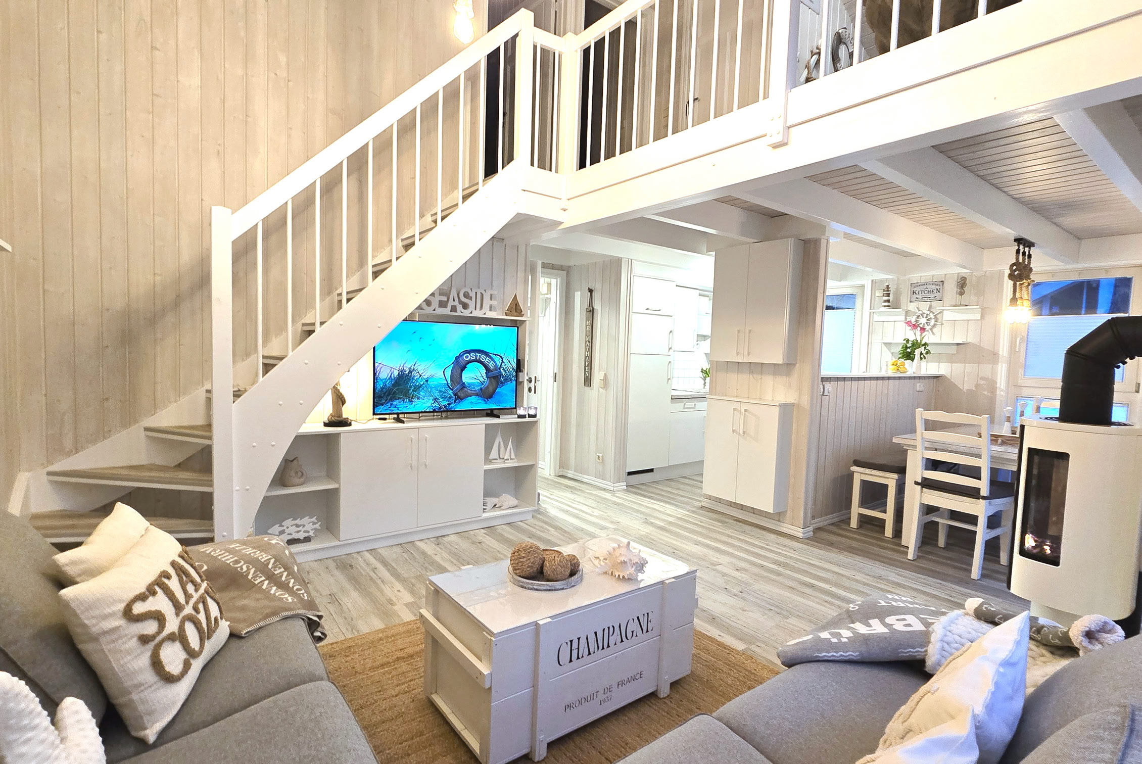 Helle Küche mit rosa Akzenten, einschließlich Toaster und Kaffeemaschine, weißen Schränken, einem Herd und einem Waschbecken. Im Hintergrund ist ein Wohnzimmer sichtbar.