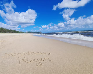 Strand mit Sand, auf dem "Rügen" geschrieben steht, Wellen im Hintergrund und blauer Himmel mit Wolken.
