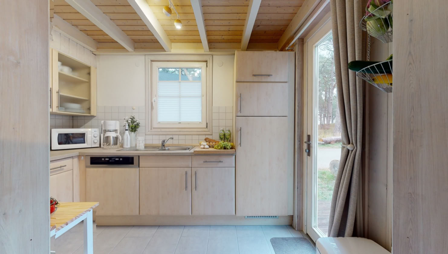 Helle Küche mit Holzschränken, einem Fenster, einem Kühlschrank, einer Mikrowelle und einer Spüle; rechts eine Glastür mit Vorhängen.