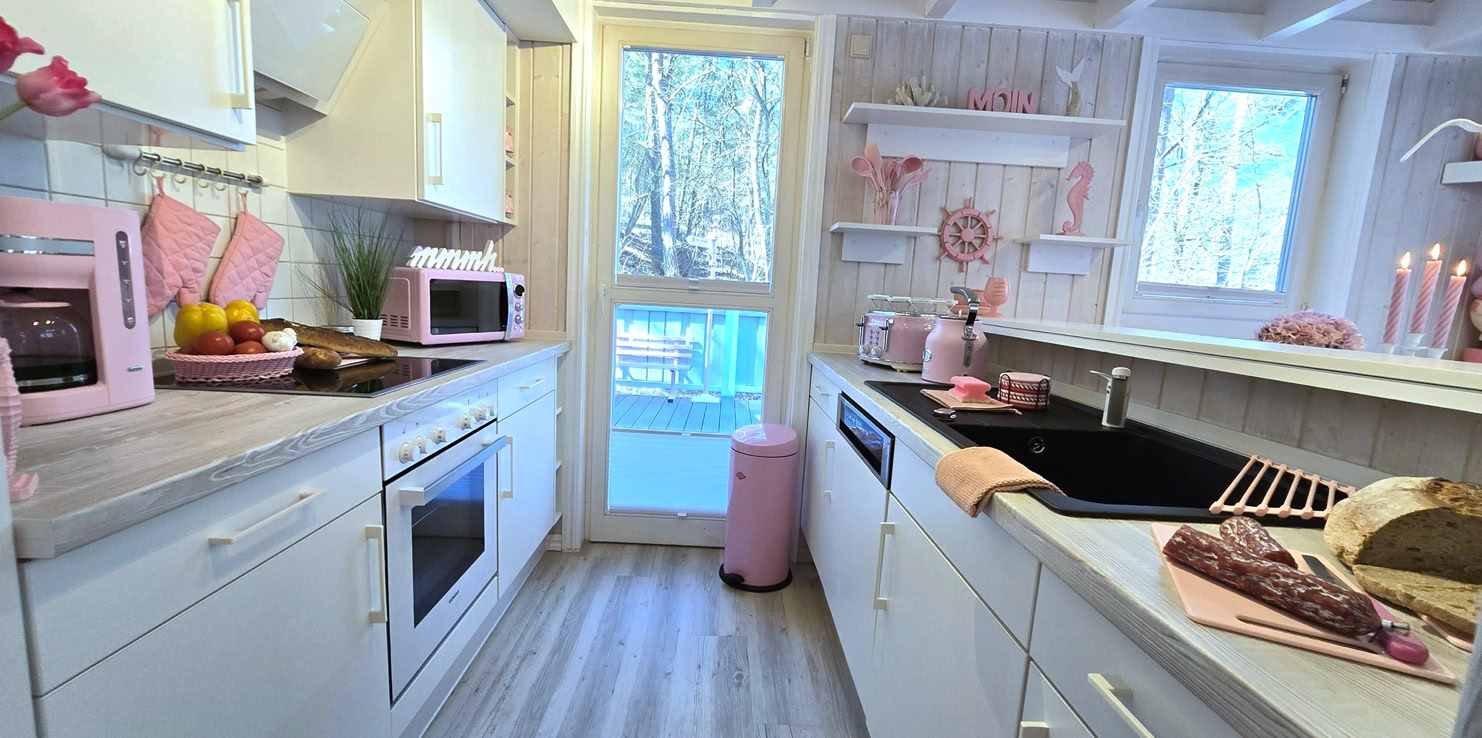 Helle Küche mit weißen Schränken, einem Herd, einer Spüle und rosa Dekorationen. Ein Fenster und eine Glastür im Hintergrund.