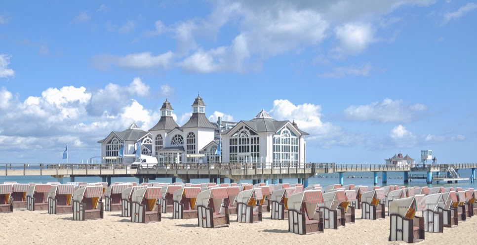 Strand mit vielen Strandkörben, im Hintergrund ein Gebäude auf einem Steg über dem Wasser, blauer Himmel mit einigen Wolken.