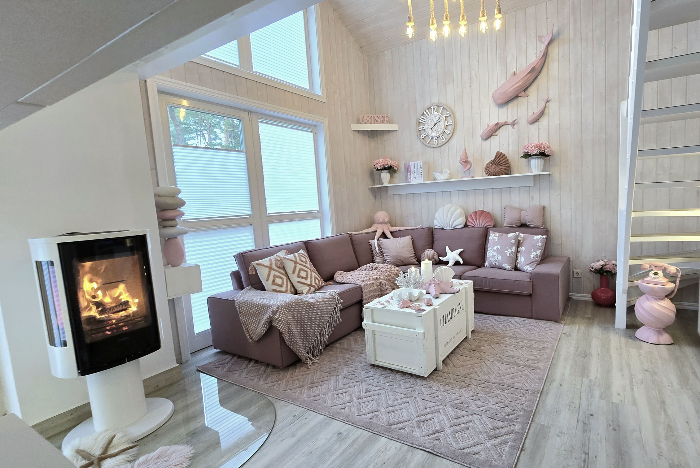 Wohnzimmer mit Kamin, Sofa, Couchtisch, dekorativen Kissen, Regalen an der Wand, Treppe und großen Fenstern.